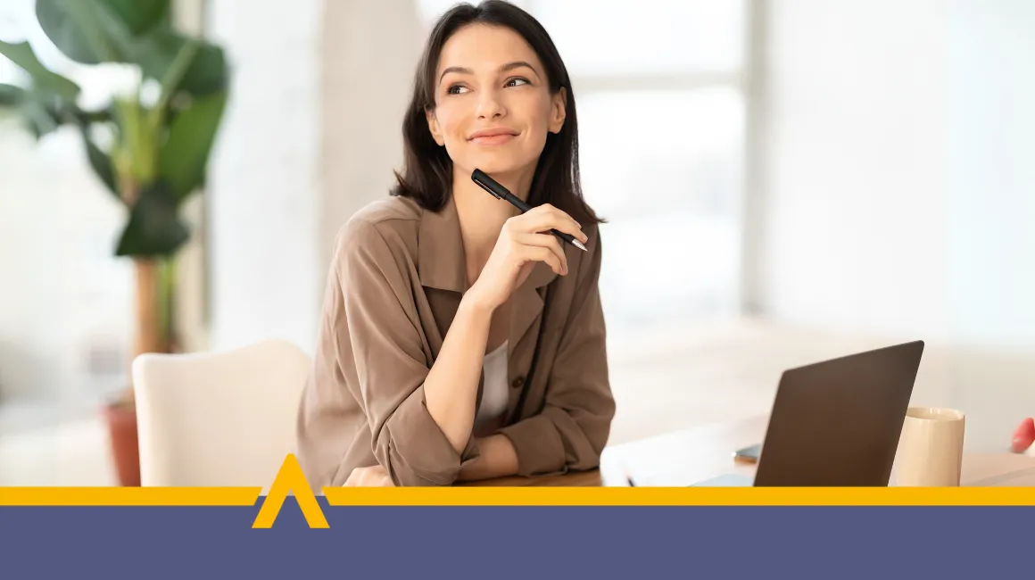 Zamyślona kobieta siedząca przy biurku z laptopem i długopisem w dłoni